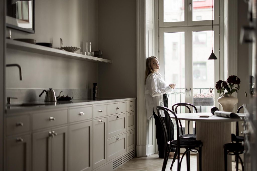 Stylish - A person stands in a modern stylish kitchen with a cup, staring out a large window. The room boasts bright cabinets and a stylish dining table with chairs.