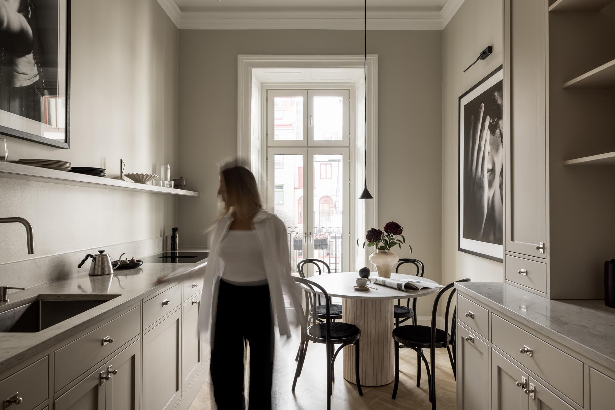 Stylish - A person walks through a stylish minimalist kitchen with beige cabinets, a large window and a cozy dining area.