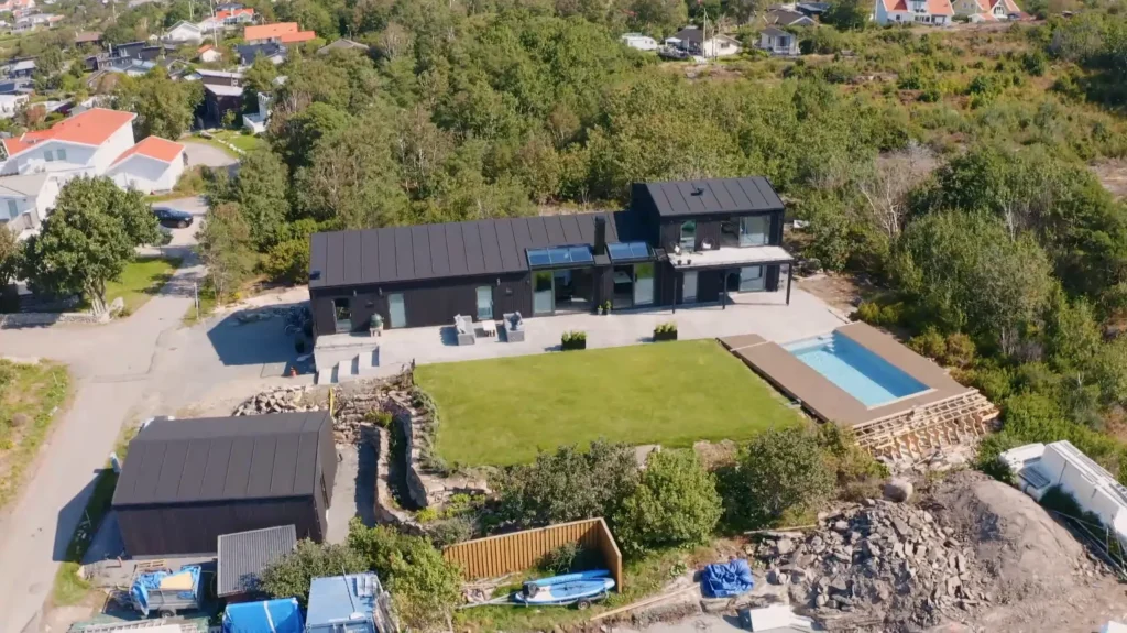 Stiligt - Flygfoto över ett modernt svart hus på Grand Designs, med en stor gräsmatta, pool och uteplats, inbäddat bland träd nära Kattegattkusten och närliggande hus.