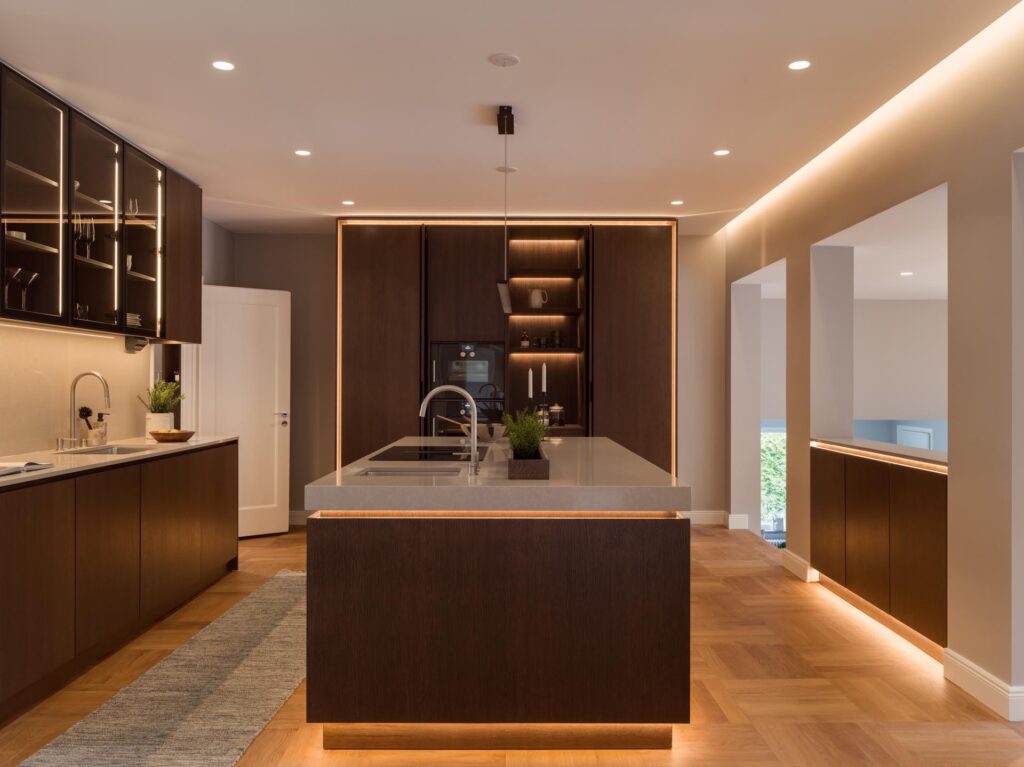Stiligt - Modern kitchen with dark wood cabinets, a central island with sink and plants, integrated appliances, under-cabinet lighting, wooden floors, and a smart wooden kitchenette with pocket doors for seamless storage.