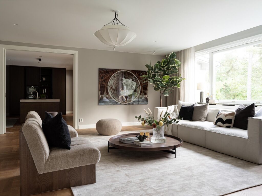 Stiligt - Modernt vardagsrum med neutralt tonade möbler, ett runt soffbord med dekor, stort fönster, väggkonst, och utsikt in i ett mörkt träkök med pocketdörrar i bakgrunden.