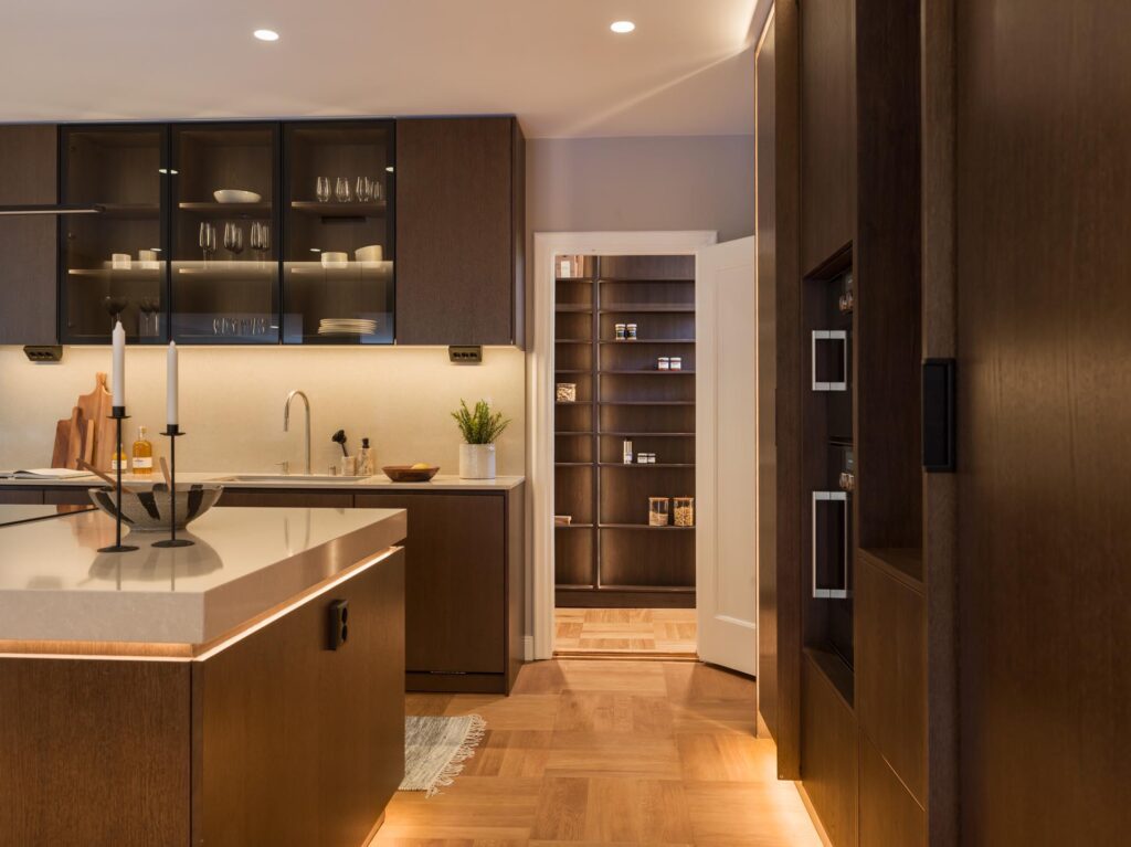 Stiligt - Modernt kök med mörka träskåp, en ljus bänkskiva, belysning under skåpet och en öppen dörr till ett skafferi – perfekt för ett träkök med pocketdörrar och smarta hyllor.