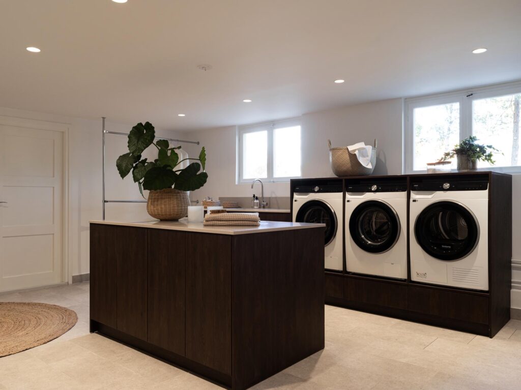 Stylish - Modern laundry room with three front-loading machines, a dark wooden island with neatly folded towels, a basket and a plant, under bright overhead lighting adjacent to large windows and a wooden kitchenette with pocket doors.