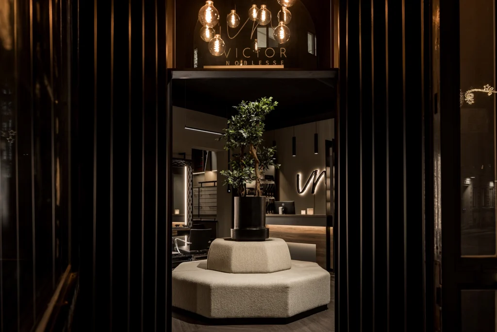 Se Stiligts unika inredning av Victor Noblesse frisörsalong – Nordens enda Wella Flagship Salon. Se detta och fler projekt för inspiration! - Stiligt inrett entréområde hos Victor Noblesse med en elegant sittmöbel och grön växt i fokus, omgiven av modern belysning och sofistikerad interiör.