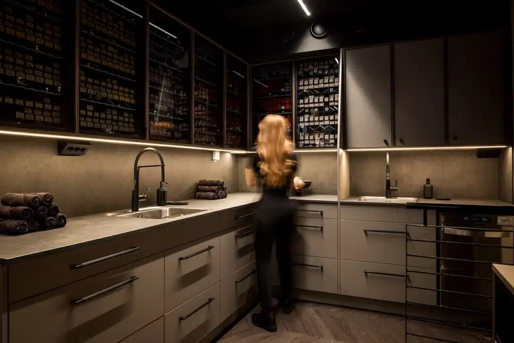 Stiligt - En kvinna med långt hår står i ett modernt, svagt upplyst kök, hennes look påminner om stilar från Victor Noblesse frisörsalong, omgiven av hyllor och skåp fyllda med burkar och flaskor.