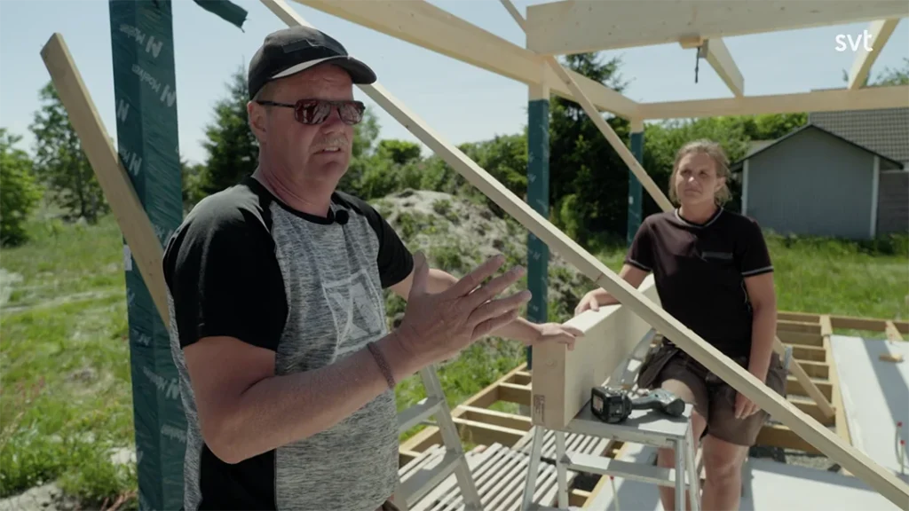 Stylish - Two people standing at a wooden house construction site in Sandby - House Dreams on a sunny day; one is talking while gesturing, and the other is standing at a workbench with tools.