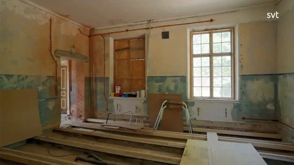 Stiligt - Ett rum under renovering med ofärdiga väggar, synliga rör, träplankor på golvet, ett fönster och byggmaterial utspridda.