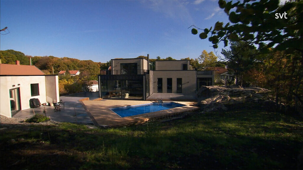 Stiligt - Modernt tvåvåningshus med stora fönster, en pool och en uteplats, omgiven av träd och grönska under en klar himmel.