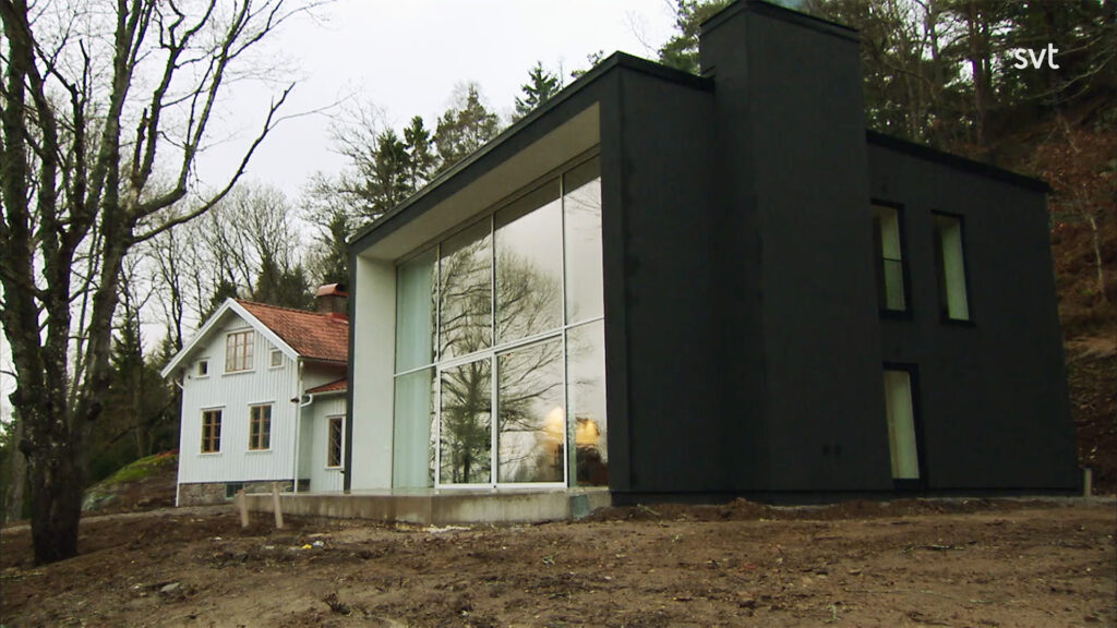Stiligt - Ett modernt svart hus med stora fönster står bredvid ett traditionellt vitt hus med rött tak, omgivet av träd på en röjd tomt.