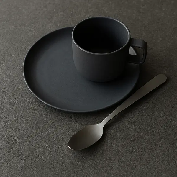 Stiligt - En mattsvart kopp står på ett matchande fat bredvid en metallsked, allt placerat på en mörk, strukturerad bänkskiva till kök för en modern kökslook.