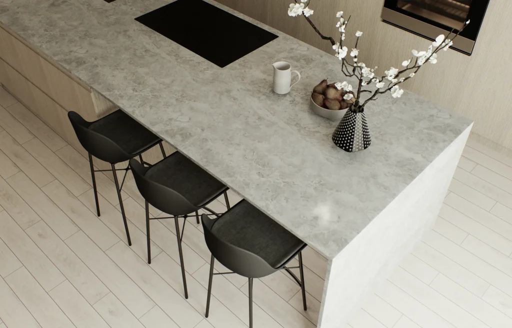 Stiligt - En bänkskiva till kök i marmor bildar köksön, omgiven av tre svarta stolar, en mugg, en skål med frukt och en vas med vita blommor på det ljusa trägolvet.